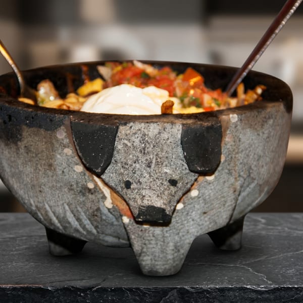 Molcajete bowl with a pig's face, filled with food, sour cream, and two spoons.